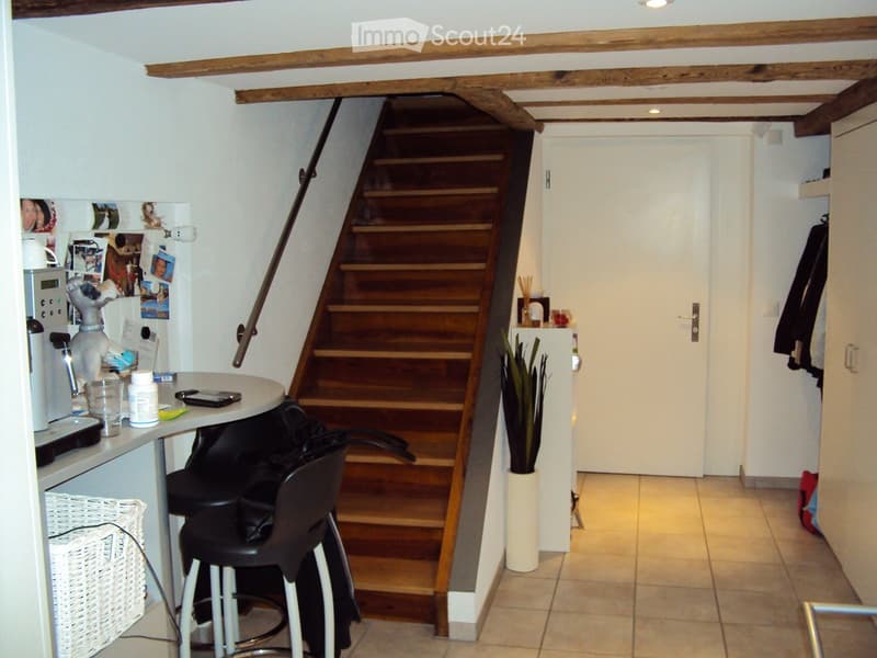 A small, cozy hallway with a wooden staircase leading upwards, featuring a small table with stools, a refrigerator with photos, and a potted plant near the stairs.