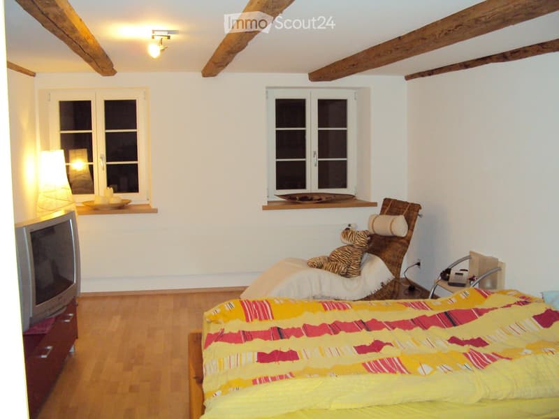 Cozy bedroom with wooden floors, exposed wooden beams, two windows, a bed with a colorful bedspread, a chair with a stuffed animal, a small table, a lamp, and a television on a stand.