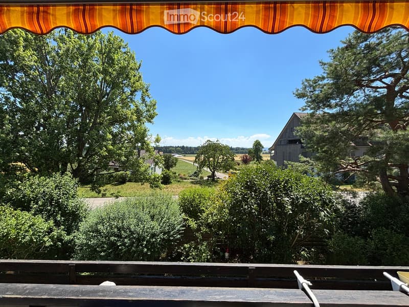 Balcony with wooden railing, striped awning, overlooking a rural landscape with trees, fields, and houses.