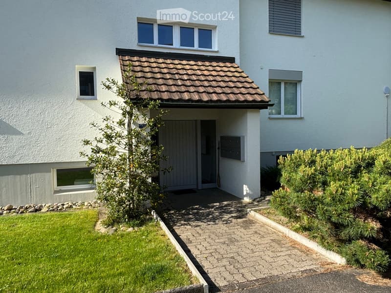 Two-story house with white exterior, tiled roof, multiple windows, and a small garden area with a pathway leading to the entrance.