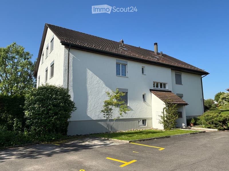 Two-story white house with a brown tiled roof, multiple windows, and a small attached garage. The house is surrounded by greenery and has a paved driveway with parking spaces.