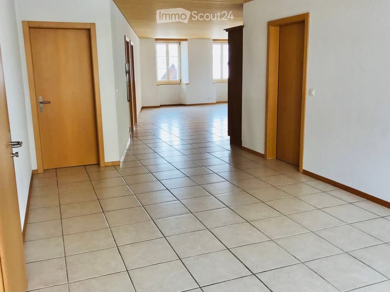 Empty hallway with tiled floor, white walls, and wooden doors on both sides. Natural light from windows at the end of the corridor.
