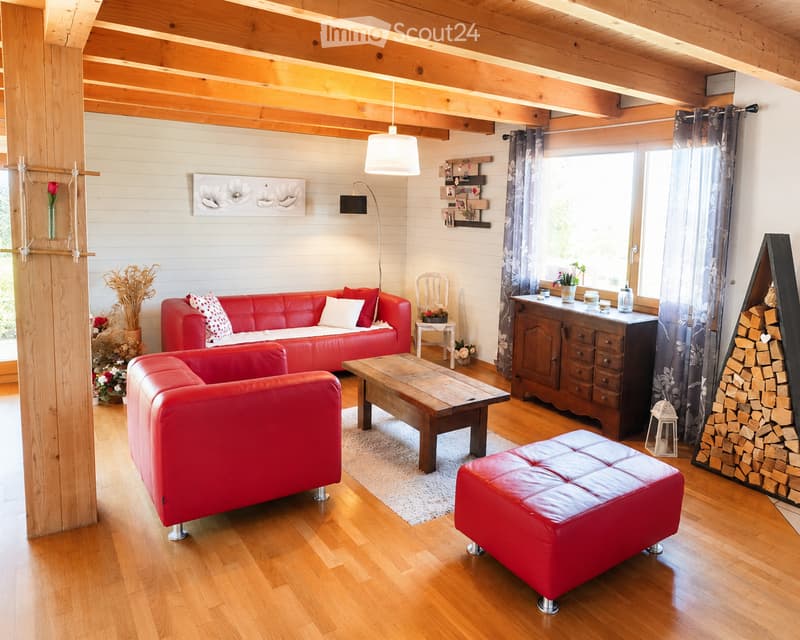Bright living room with wooden beams, red leather sofa, coffee table, ottoman, and a wooden cabinet. Large windows with curtains, decorative items, and a stack of firewood.