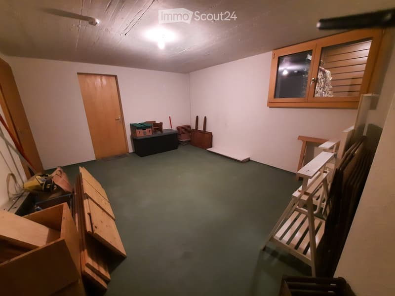 Empty room with bare walls, green carpet, and a wooden door. Various construction materials and tools are scattered around, including wooden planks, a ladder, and storage boxes.