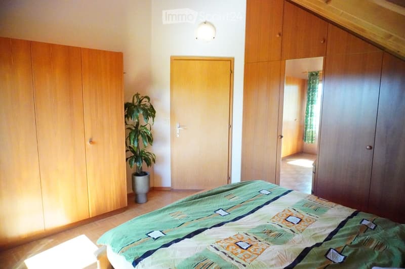 Bright bedroom with wooden floor, large wardrobe, bed with green and orange patterned duvet, potted plant, and a mirror on the wardrobe.
