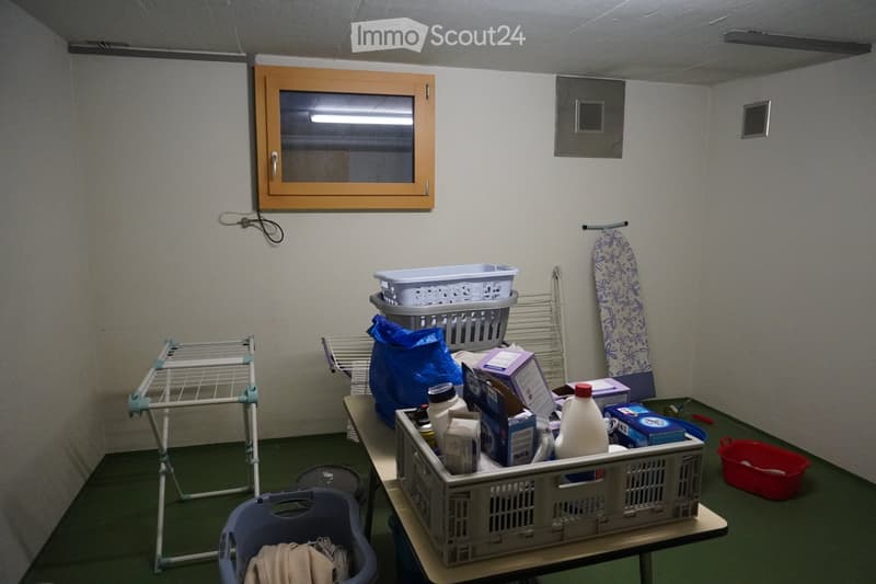 A small utility room with a table holding laundry supplies, a drying rack, and an ironing board against the wall.