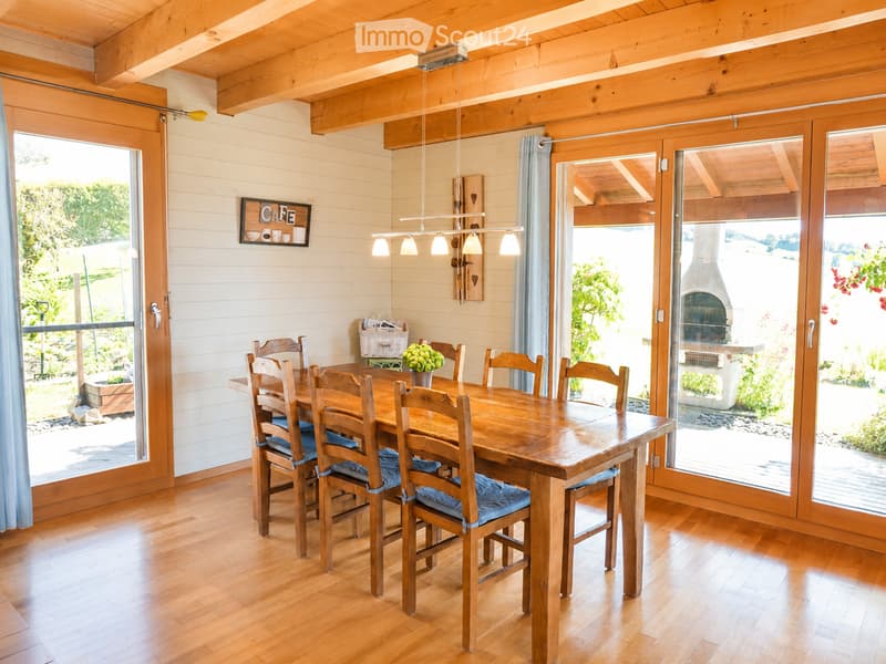 Bright dining area with wooden dining table and chairs, large windows, exposed wooden beams, and a decorative sign on the wall.