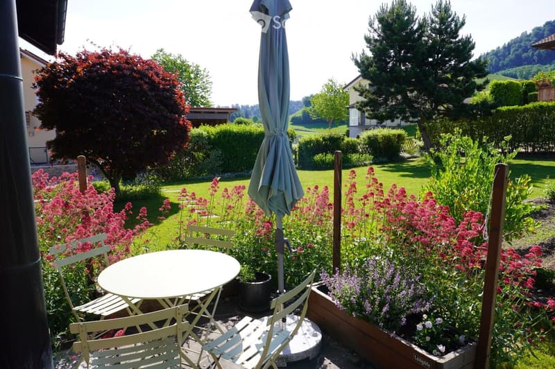Garden area with a round table, four chairs, and an umbrella, surrounded by colorful flowers and greenery, with a well-maintained lawn and trees in the background.