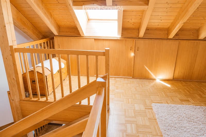 Spacious attic room with wooden beams, exposed wooden staircase, built-in wooden cabinets, and a small wooden chest. The floor is covered with herringbone-patterned wooden planks and a white rug is placed near the staircase.