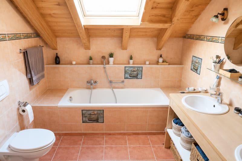 Bathroom with a bathtub, toilet, sink, and mirror. Features wooden beams on the ceiling, tiled walls, and decorative items including vases and picture frames.