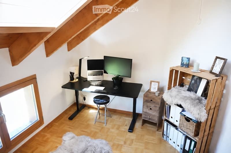 Cozy attic workspace with wooden slanted ceilings, a black standing desk, a stool, a laptop, a monitor, a small plant, and a wooden shelf with books, a furry throw, and decorative items.
