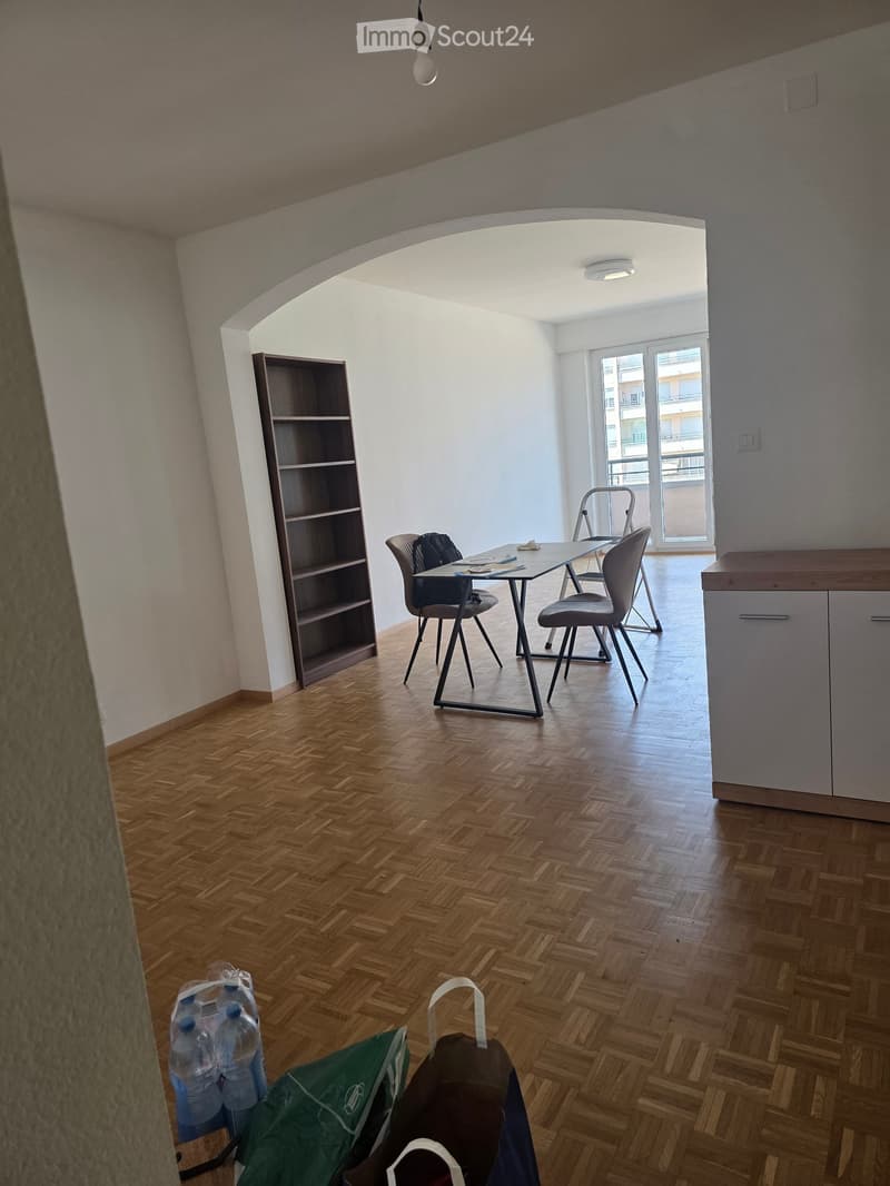 A modern, unfurnished room with wooden flooring, a table, chairs, and a shelf. The room has large windows and a balcony view.