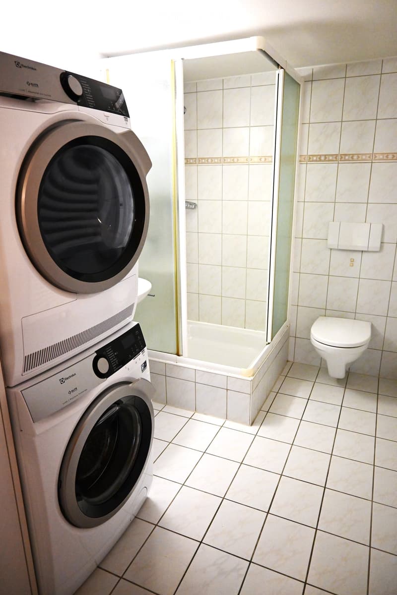 A bathroom with a white tiled floor, a white toilet, a glass shower enclosure, and a white washing machine and dryer stacked on top of each other.