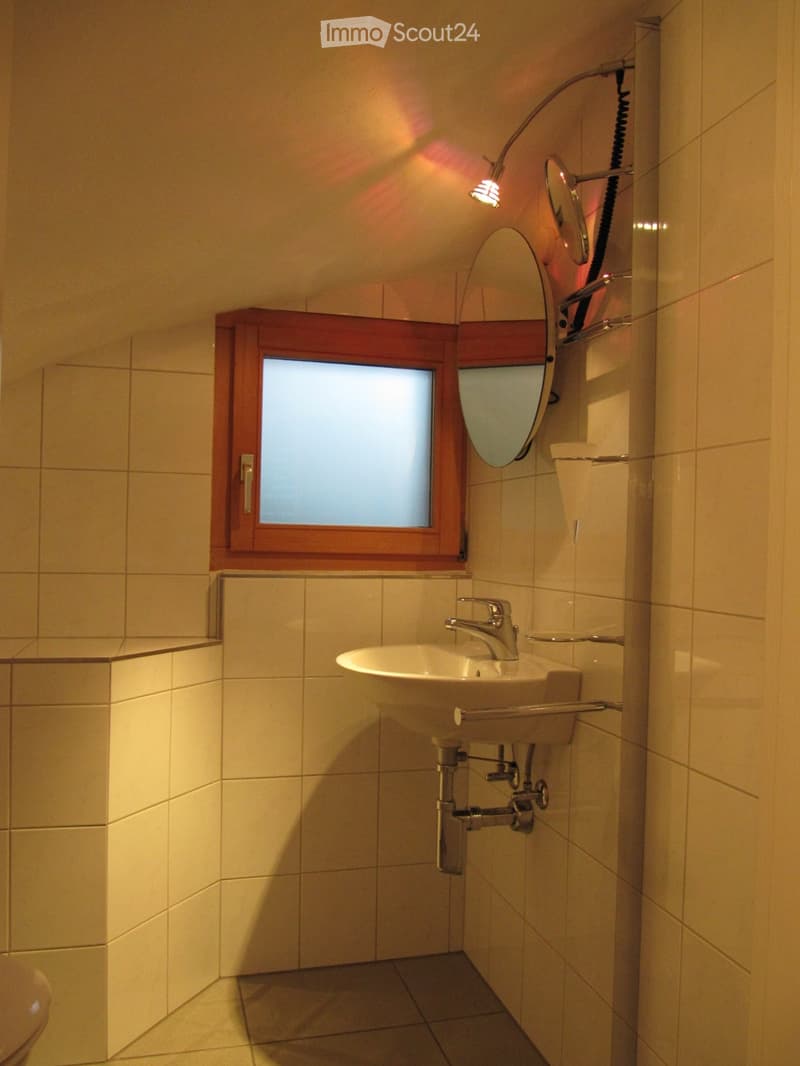 Modern bathroom with a white sink, chrome faucet, round mirror, and a small window with wooden frame. Light-colored tiles on walls and floor.