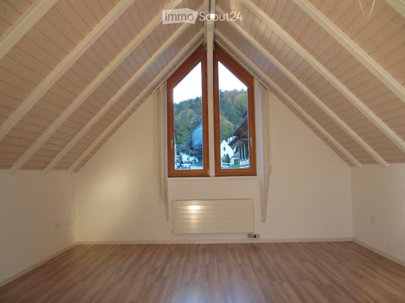 Empty attic room with wooden floors, white walls, and large windows offering a view of the surrounding area.
