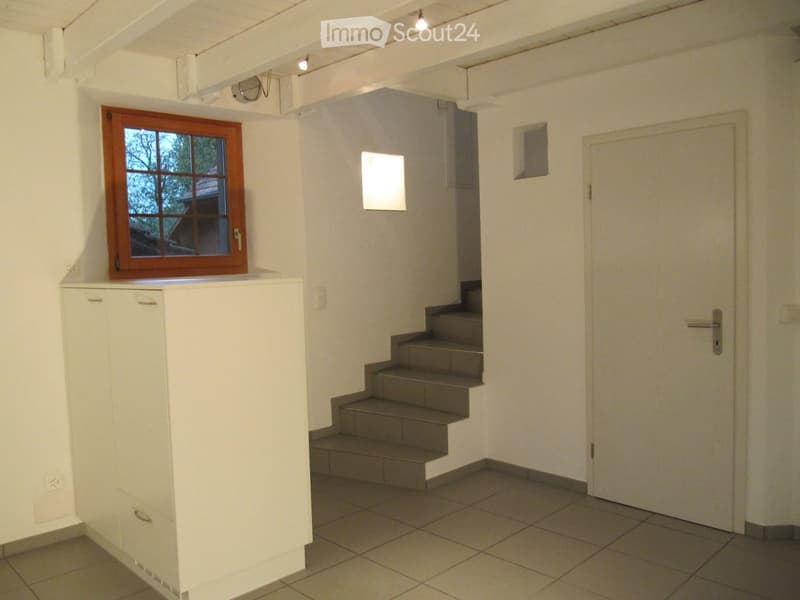 Empty room with tiled floor, white walls, a staircase leading upwards, a window with wooden frame, and a white cabinet with drawers.