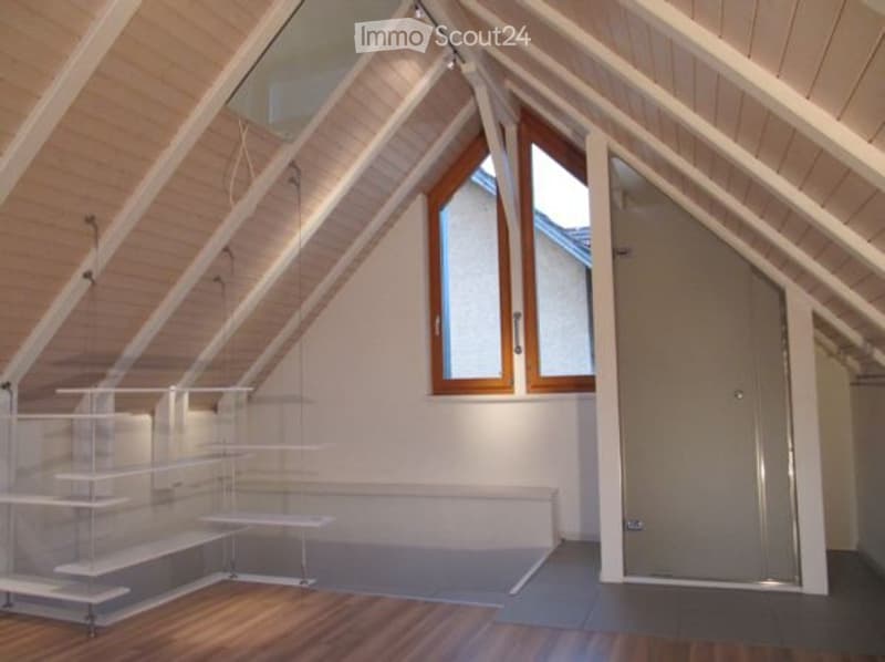 Bright, airy attic space with wooden beams, exposed ceiling, large windows, and built-in shelving units.