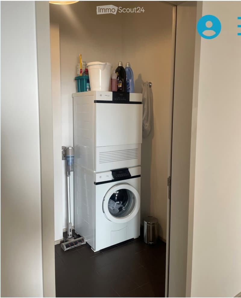 Compact bathroom with a stacked washing machine and dryer, cleaning supplies on top, a towel hanging on the wall, and a trash can on the floor.