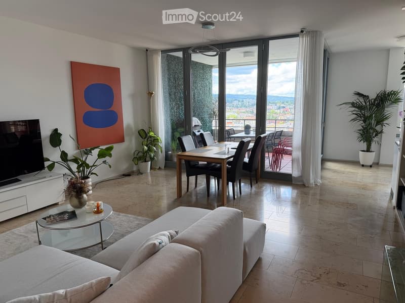 Modern living and dining area with a light grey sofa, glass coffee table, dining table with chairs, large windows with a view, and decorative plants.
