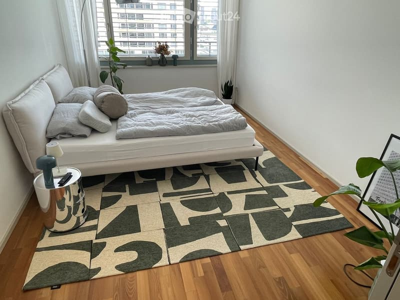 Modern bedroom with a white platform bed, nightstand, side table, and decorative plants. The room features a geometric patterned rug and large windows with city views.