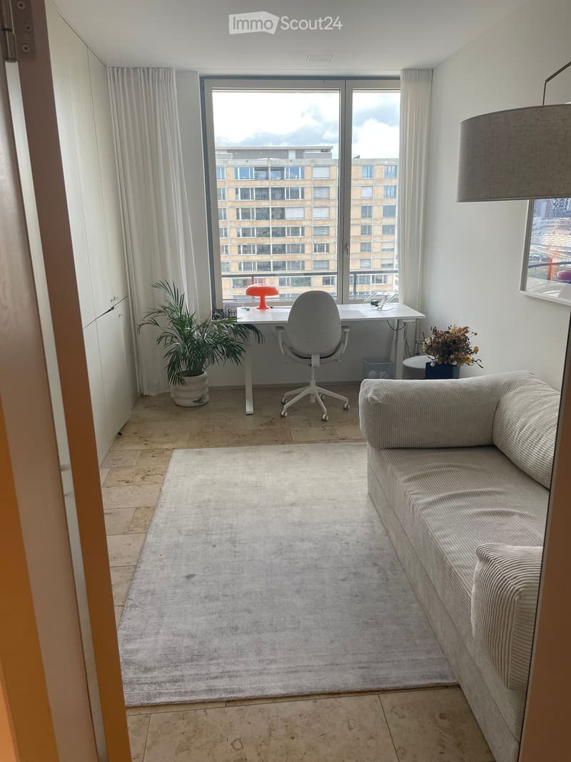 Modern living room with a sofa, chair, table, lamp, plant, and flower arrangement. Large windows offer a view of the city.