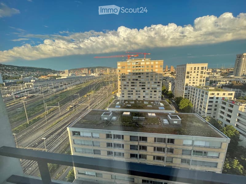 View from a balcony showing a cityscape with multiple residential buildings, train tracks, and a partly cloudy sky.