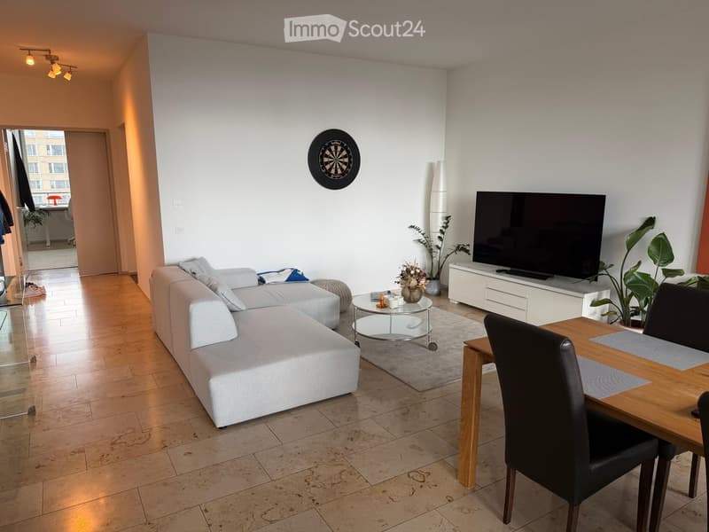 Modern living room with a grey sectional sofa, coffee table, dining table with chairs, TV, and plants. The room features wooden flooring and white walls.