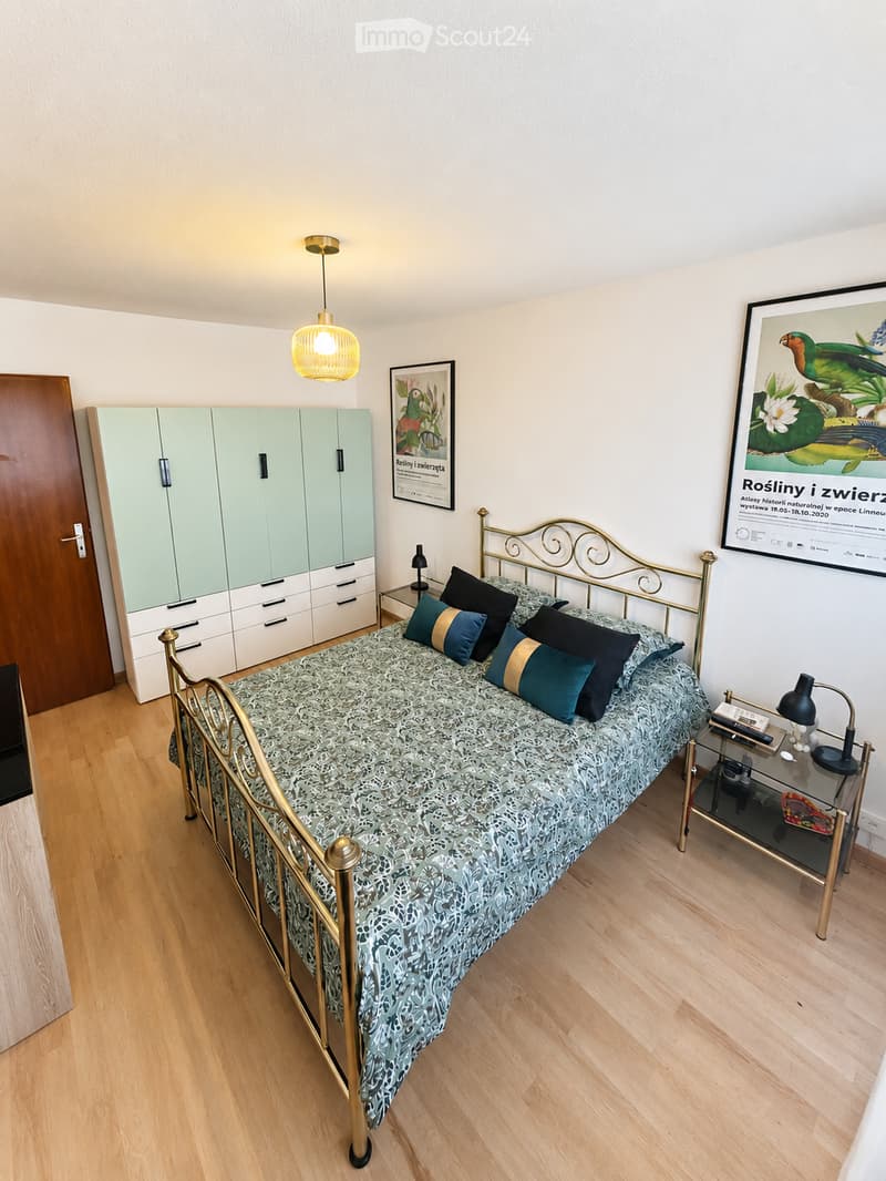Cozy bedroom with a wooden floor, a metal-framed bed with patterned bedding, nightstand with lamp, wardrobe, and artwork on the walls.