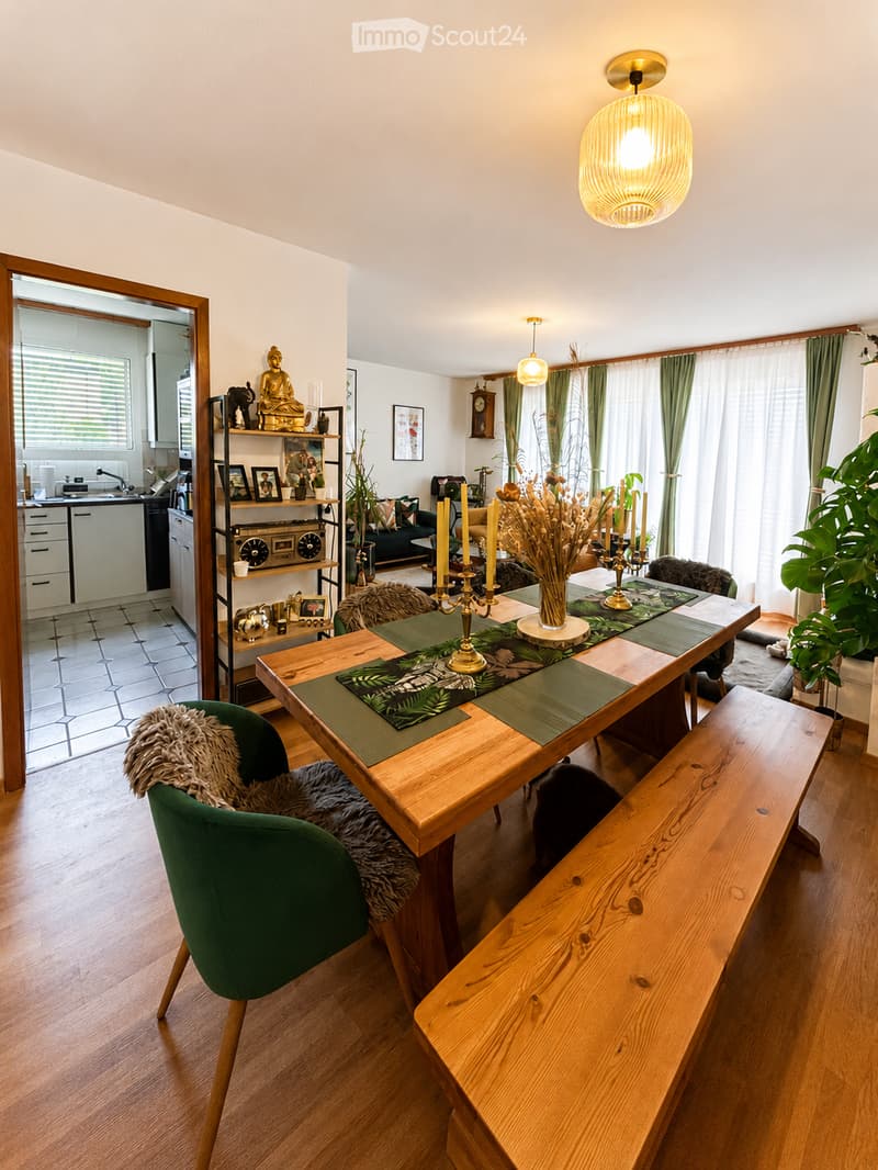 A well-lit dining area with a wooden table, green placemats, and a bench. A green chair with a fur throw is visible. A shelf with decorative items and a framed picture is in the background, along with a kitchen area visible through an open doorway.