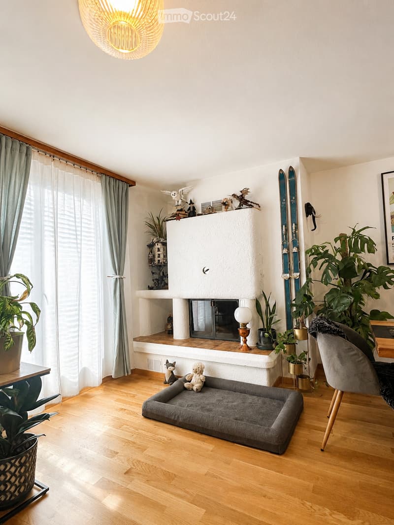 A cozy living room with wooden flooring, a fireplace, a dog bed, a sofa, a table, a chair, and decorative plants.
