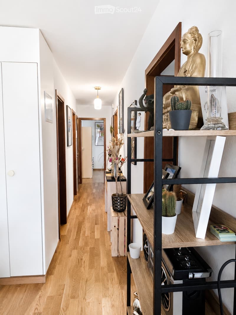 A modern hallway with light wooden flooring, white walls, and several doors leading to different rooms. A tall black shelving unit holds decorative items, including a golden statue, potted plants, and framed pictures. A small table with a vase of dried fl