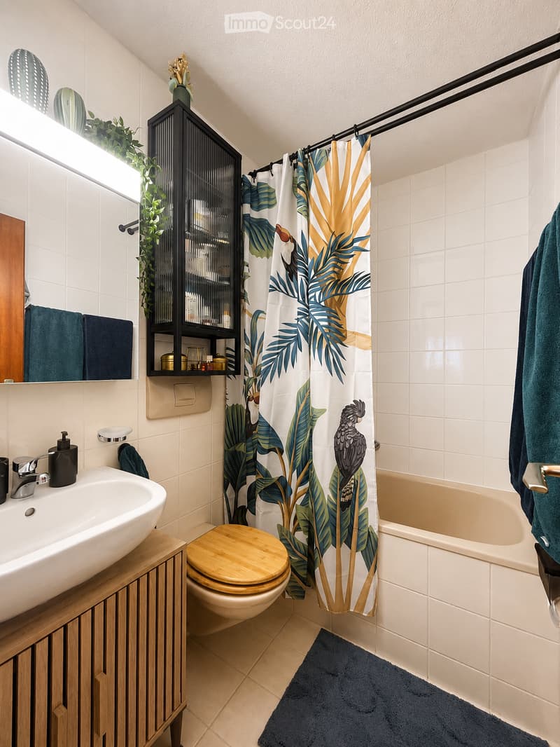 Modern bathroom with white tiles, a wooden toilet seat, a sink with a faucet, a shower curtain with a tropical design, a dark blue towel, and a small plant on the windowsill.