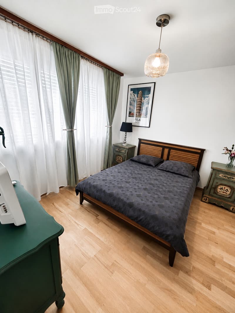 Cozy bedroom with wooden flooring, a double bed with dark bedding, two nightstands with lamps, a framed picture on the wall, and a hanging pendant light.