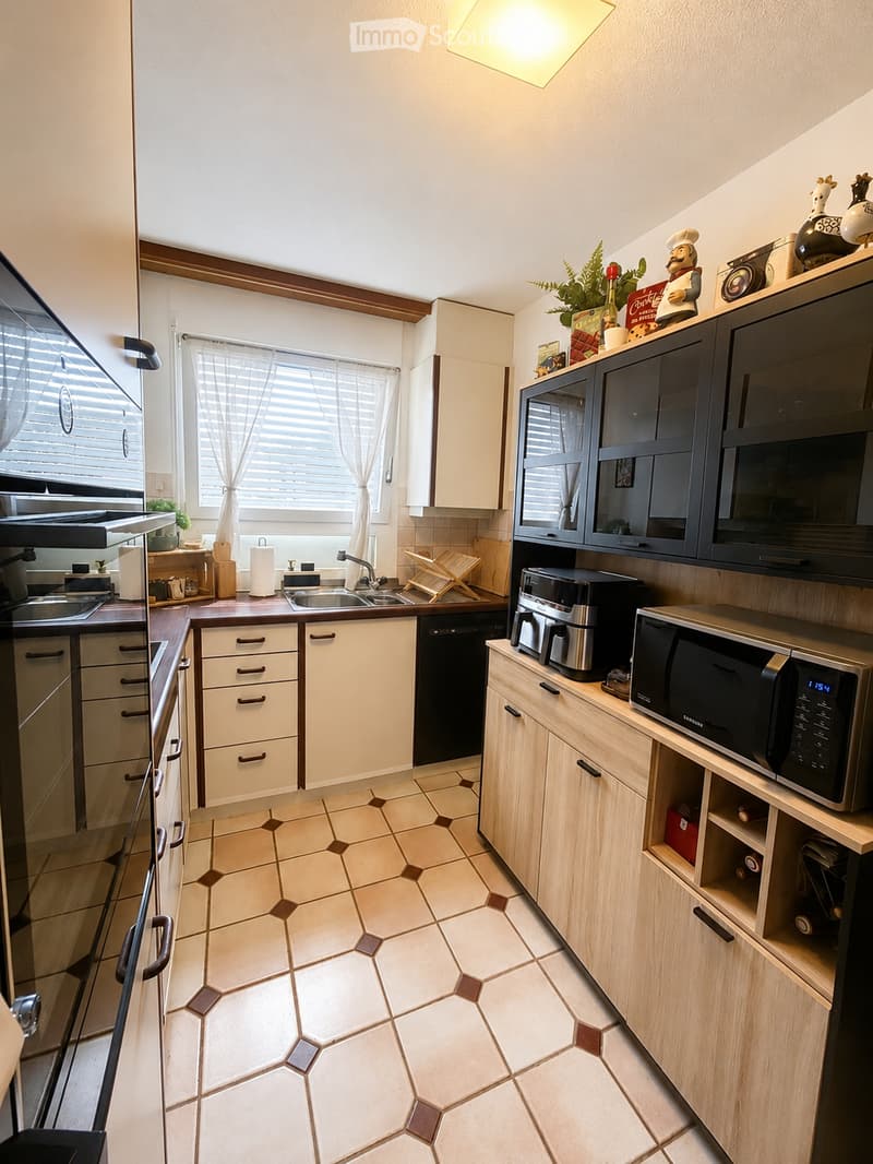 Modern kitchen with white cabinets, wooden countertops, and a tiled floor. Features include a sink, microwave, dishwasher, coffee maker, and various kitchen appliances.