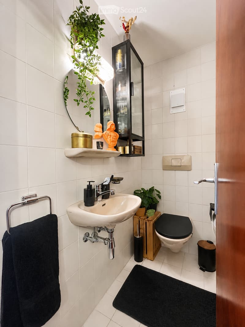 Modern bathroom with white tiles, a sink, toilet, mirror, and decorative items including plants and a small wooden shelf.