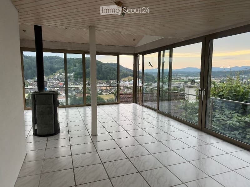 Bright, open space with large glass windows offering a scenic view of the valley and mountains. The room features a tiled floor and a wood-burning stove, with a wooden ceiling and modern lighting fixtures.