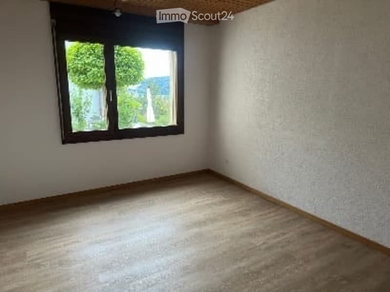Empty room with wooden flooring and white walls, featuring a large window with a view of greenery outside.