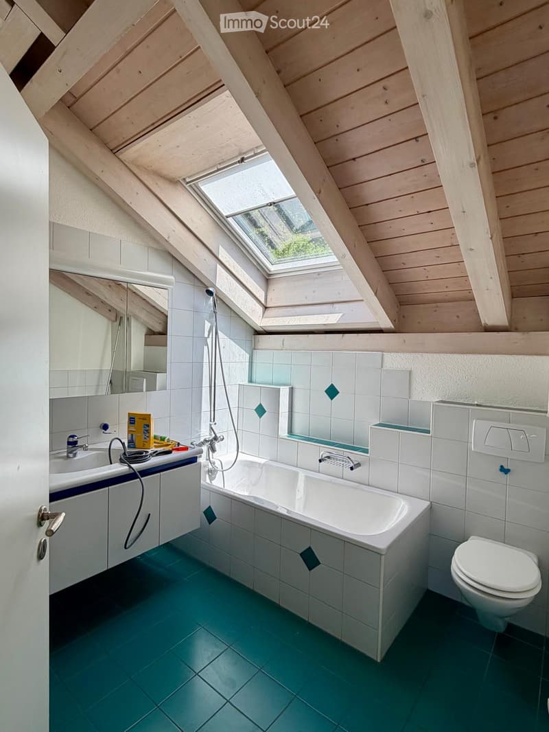 Bathroom with a bathtub, toilet, sink, and showerhead. The room features a skylight window, wooden slanted ceiling, and green tiled floor.