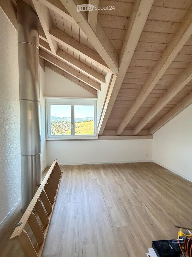 Empty attic space with exposed wooden beams, wooden flooring, and a large window offering a scenic view.