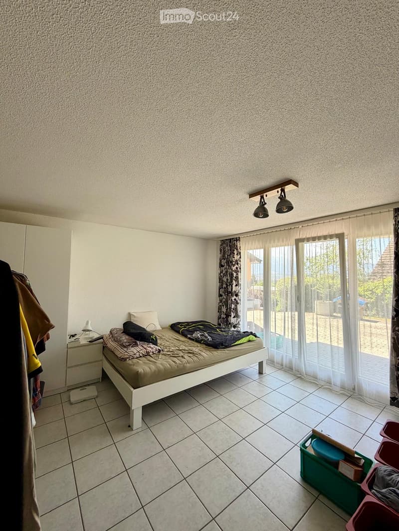 Bedroom with tiled floor, white walls, large window with curtain, bed with mattress and pillows, nightstand with lamp, wardrobe, and storage bins.