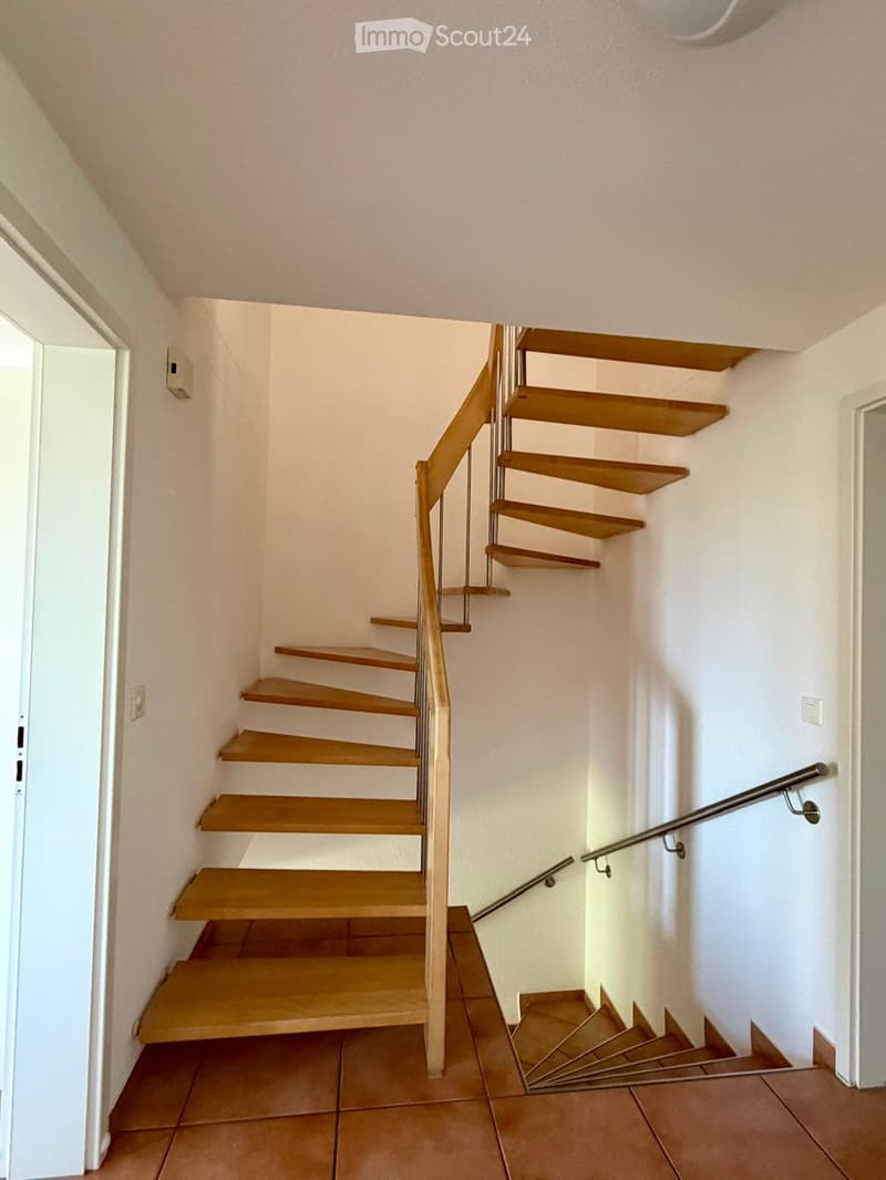 Spiral staircase with wooden steps and metal railing, leading to an upper level. The walls are white, and the floor is tiled.