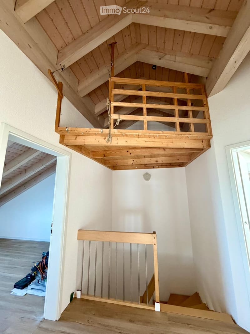 A modern hallway with exposed wooden beams and a staircase leading to an upper level. The staircase features a wooden railing with metal balusters and a rope handrail.