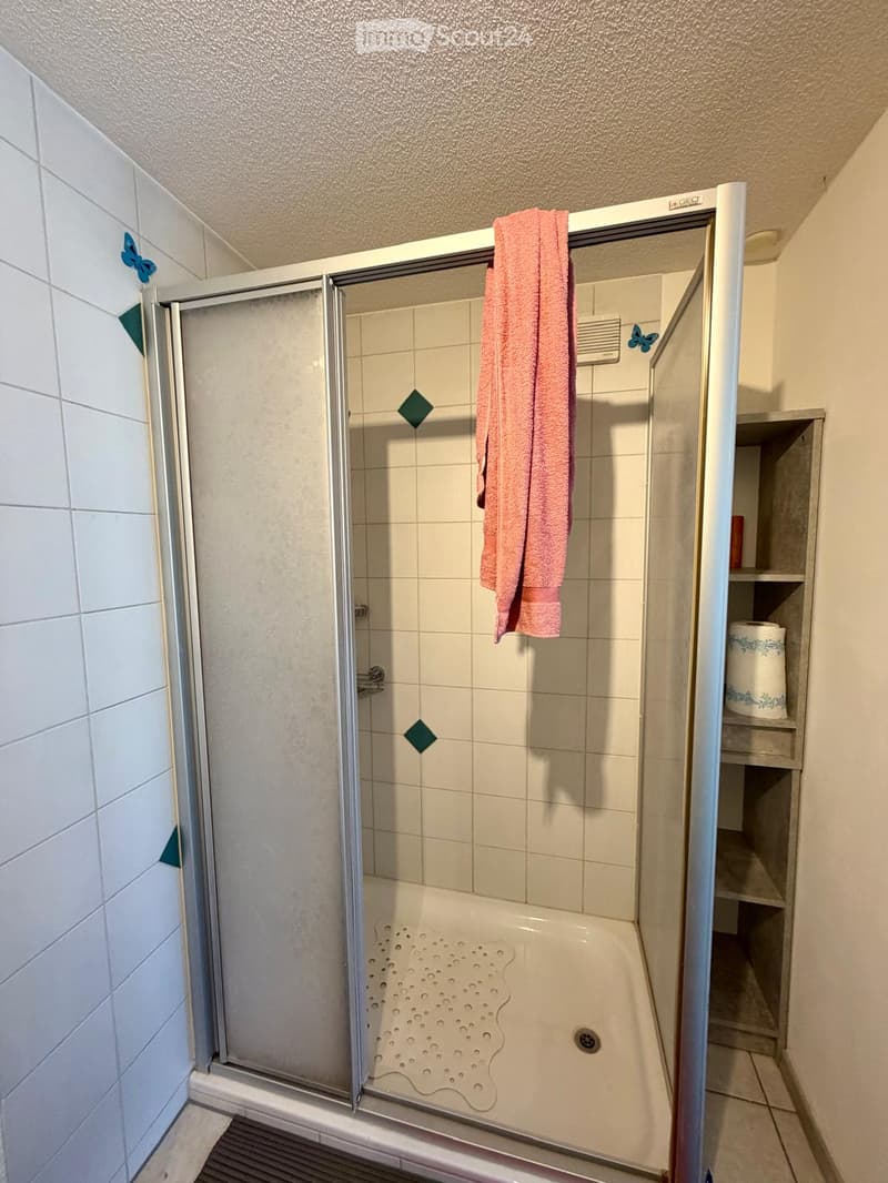 A modern bathroom with a walk-in shower, tiled walls, a towel hanging on the glass door, and a shelf with toilet paper.