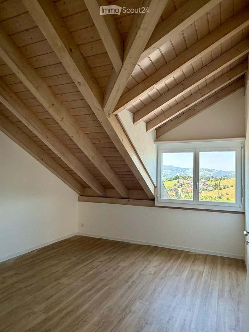Empty attic room with wooden slanted ceilings, wooden flooring, and a large window offering a scenic view of the countryside.