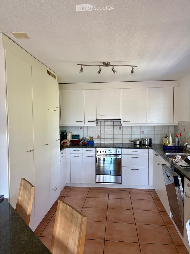 Modern kitchen with white cabinets, stainless steel appliances, tiled backsplash, and wooden chair visible.