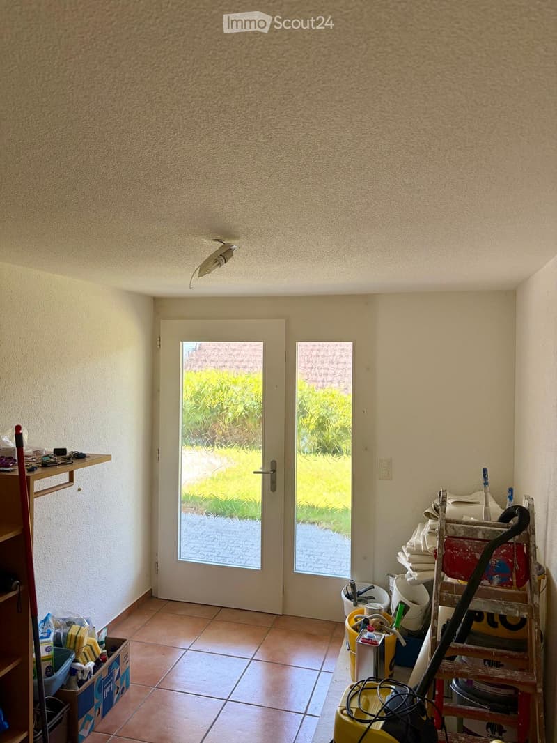 A small, tiled room with two doors leading outside, a shelf on the left, and cleaning supplies and equipment on the right.