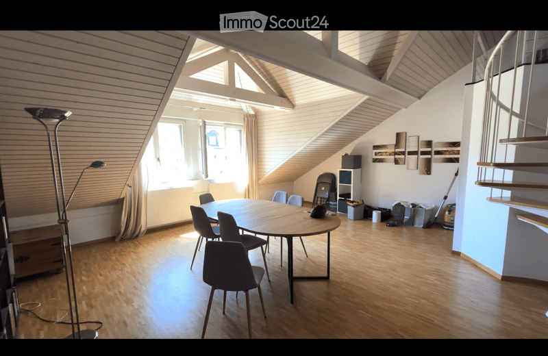 Bright, open room with wooden floors and slanted ceilings, featuring a round wooden table surrounded by chairs, a tall floor lamp, and a shelf with various items.