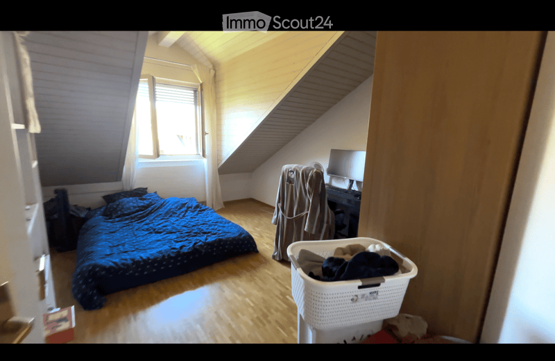 Bedroom with slanted wooden ceiling, bed with blue bedding, desk with computer, laundry basket, and clothing rack.
