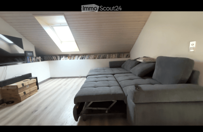Modern living room with a large sectional sofa, bookshelf, and a TV screen. The room features wooden flooring and a slanted ceiling with skylights.