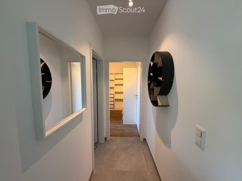 Modern hallway with white walls, tiled floor, and recessed lighting. Features a large mirror on the left and a decorative wall-mounted sculpture on the right, leading to an open doorway.